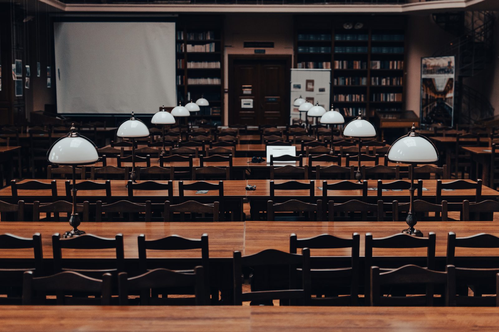 A university reading hall: long tables, green-shaded lamps and shelved stacks receding towards the far wall.
