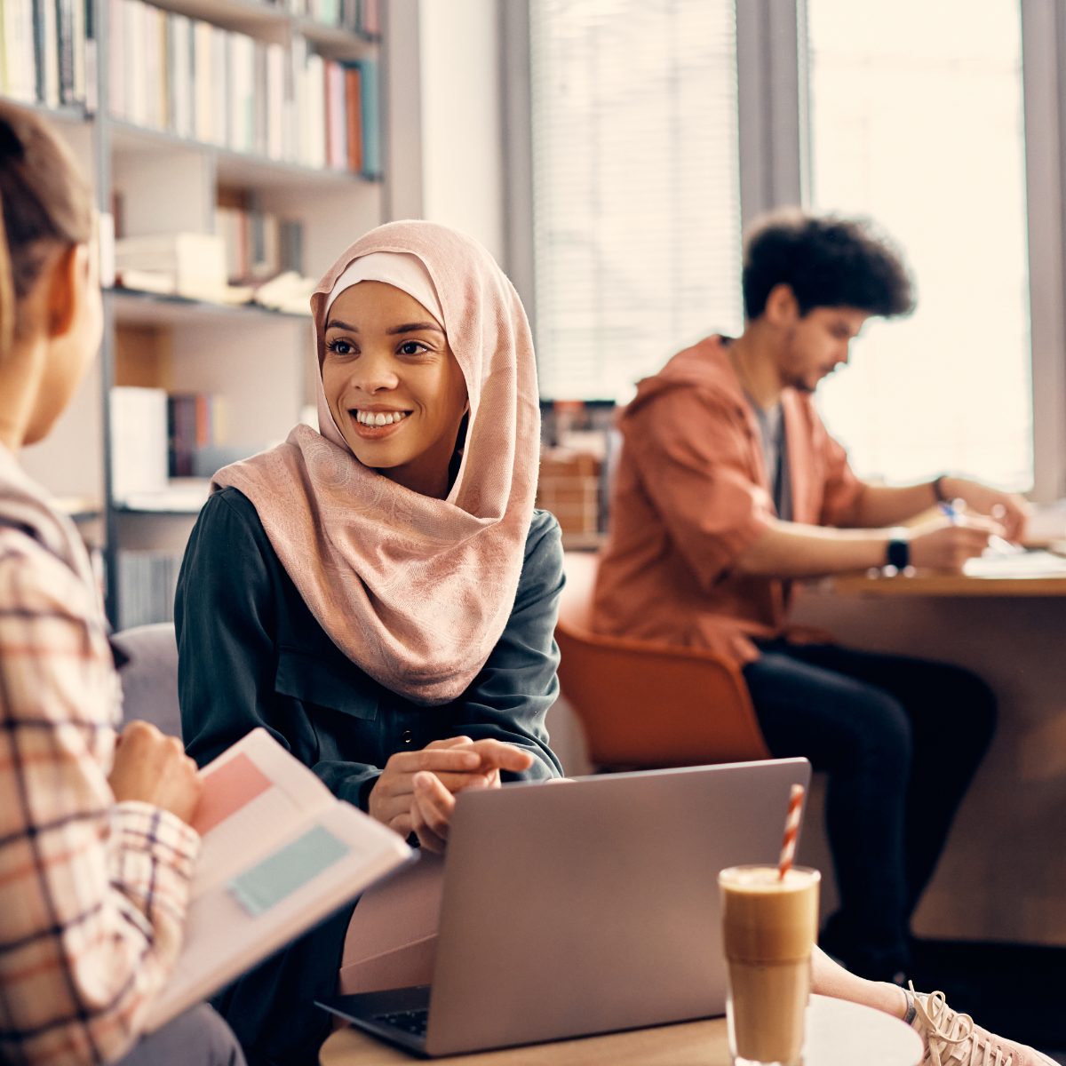 طالبتان في حوار حول حاسوب محمول داخل مكتبة، وخلفهما طالب يقرأ على طاولة.
