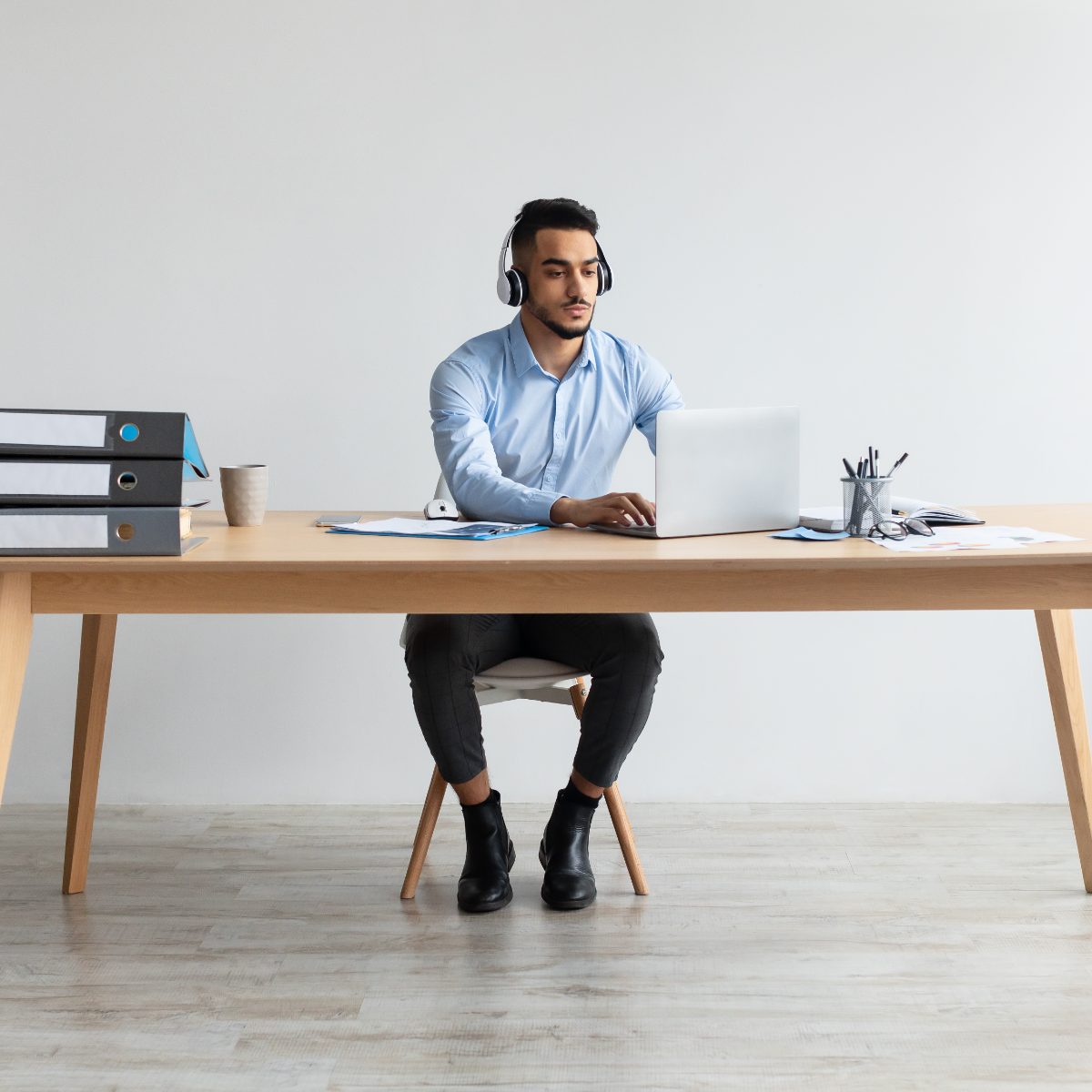 A learner in a collared shirt working at a laptop with headphones, files and papers laid out on the desk beside him.